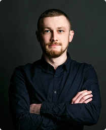 Een man met een kortgeknipte baard en blauwe ogen staat tegen een donkere achtergrond, gekleed in een donker shirt en met zijn armen over elkaar, en straalt zelfvertrouwen uit.