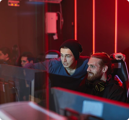 Twee mannen in een slecht verlichte speelzaal staren geconcentreerd naar een computerscherm. De ene wijst naar het scherm, terwijl ze allebei helemaal in het spel zitten en enthousiast lijken. Rode neonlichten zorgen voor een levendige sfeer.