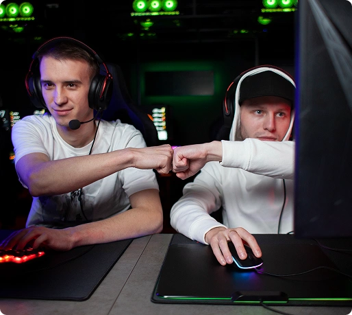 Twee gamers met een headset op geven elkaar een high five terwijl ze achter computers met kleurrijke toetsenborden zitten, wat een sfeer van teamwork en opwinding uitstraalt in een schemerige gameroom.