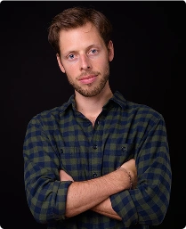 Een man met lichtbruin haar en een baard staat voor een zwarte achtergrond. Hij draagt een groen-blauw geruit overhemd, heeft zijn armen over elkaar en straalt zelfvertrouwen uit.